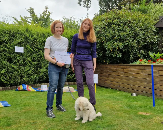 Katya outdoors with a happy client and their young poodle puppy holding a training certificate